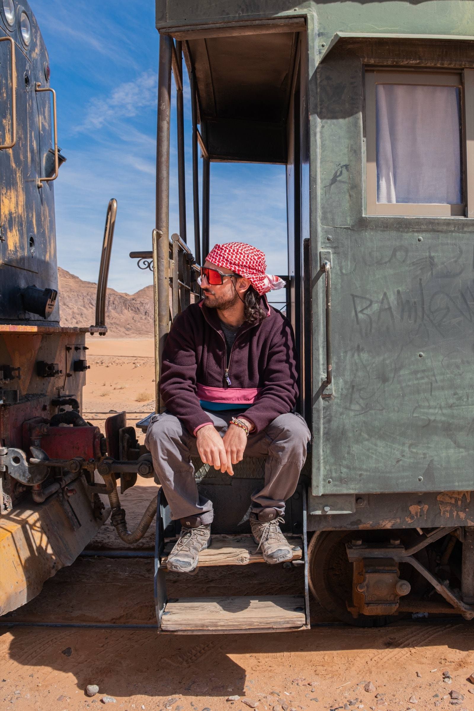 Un viaggiatore con la kefiah rossa seduto sulla scaletta di un vecchio vagone nel deserto giordano